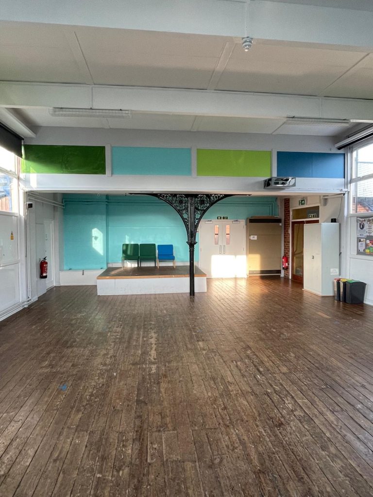 Empty indoor hall with a wooden floor, a small raised stage area, and colourful wall panels, lit by natural light from windows.