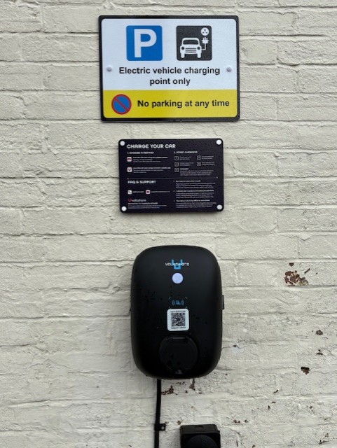 Wall-mounted electric vehicle charger with cable, beneath signs indicating “Electric vehicle charging only” and “No parking at any time,” plus an instruction panel above.