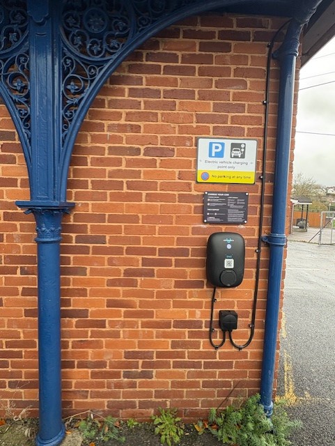 Wall-mounted electric vehicle charger with cable on a brick wall beneath signs indicating “Electric vehicle charging only” and “No parking at any time,” with an instruction panel above.