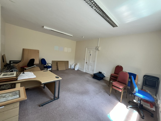 Large office room containing desks, chairs and stored furniture, showing the entrance door and carpeted floor.
