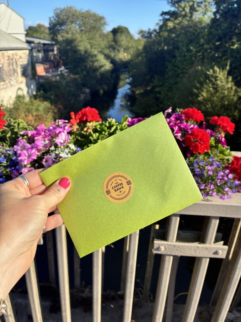 A green envelope displaying The Coffee Bank logo is held above a bridge decorated with colourful flowers. The River Otter and surrounding trees can be seen in the background.