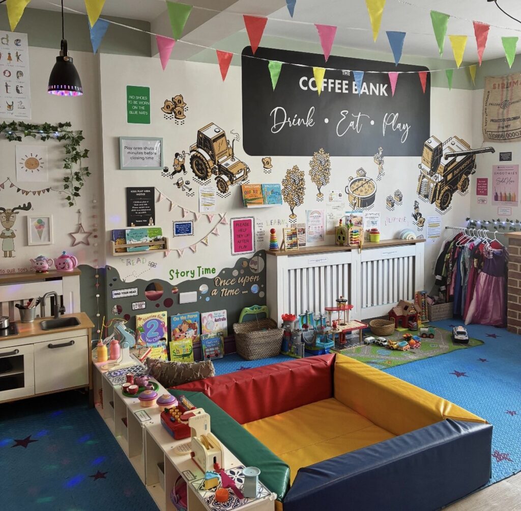 A dedicated children's play area inside The Coffee Bank, featuring toys, books, play mats, dressing-up clothes and soft play equipment. Colourful bunting hangs from the ceiling and a wall graphic displays The Coffee Bank's slogan, “Drink • Eat • Play”.