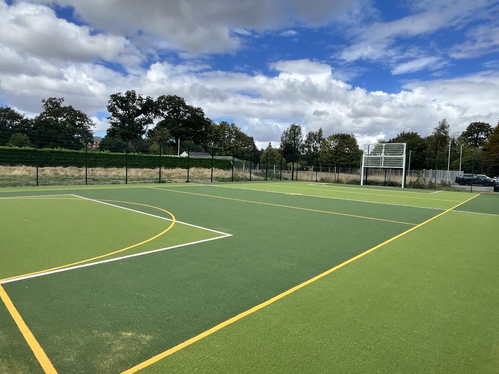 An outdoor multi-use games area with artificial turf, marked court lines and sports goals enclosed by perimeter fencing. Trees and open grassland surround the facility under a partly cloudy sky.