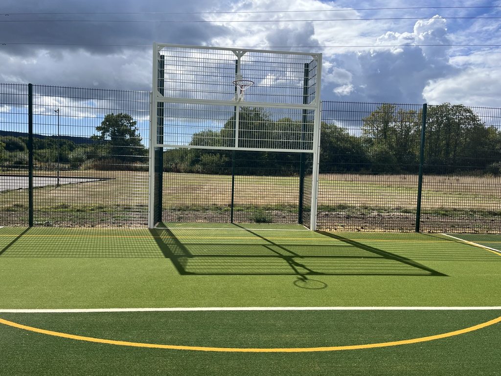 A netball hoop mounted on a fenced structure within an outdoor multi-use games area with artificial turf and marked sports court lines. Open grassland and trees are visible beyond the perimeter fencing.
